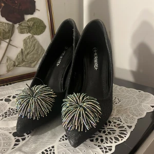 Glittriga svarta klackar - Svarta pumps från Altramarea med glittrig yta och spetsig tå. Skorna har hög klack och är dekorerade med stora pärlbroderade detaljer framtill som ger en riktigt festlig och perfekt för nyårsafton