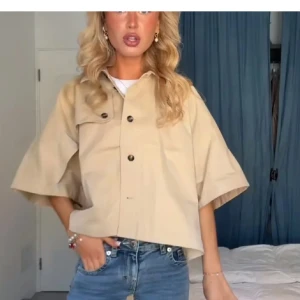 Beige overshirt med vida ärmar - Snygg beige overshirt med vida, korta ärmar och svarta knappar framtill. Jackan har en bröstficka med lock och är lätt boxig i modellen. Perfekt att slänga över en t-shirt för en chill look.