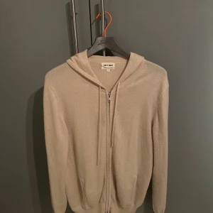 Beige kashmirhoodie från Soft Goat - Mysig beige hoodie från Soft Goat i 100% kashmir. Tröjan har dragkedja framtill, huva med snörning och två fickor. Perfekt för dig som vill ha en stilren och lyxig look med maximal komfort. Passar bäst på folk som är 180 cm ungefär