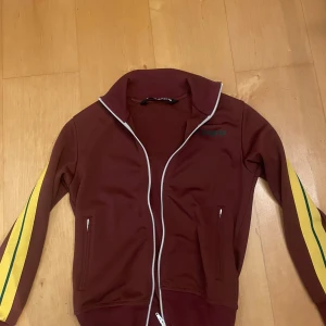 Vinröd track jacket från Palm Angels XS - Säljer en vinröd track jacket från Palm Angels med vita dragkedjor och gula ränder med grön detalj längs ärmarna. Tröjan har hög krage, två fickor med dragkedja och logga på bröstet. Tillverkad i 100% polyester för en sportig vibe.