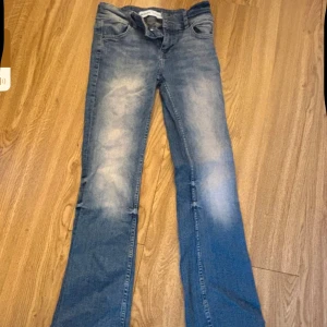 Blå bootcut lågmidjade jeans från Bershka - Snygga blå jeans från Bershka i bootcut-modell med klassisk femficksdesign. Jeansen har ljusa slitningar framtill och baktill för en cool, tvättad look. Tillverkade i jeansmaterial och passar dig som gillar lite bredare ben.