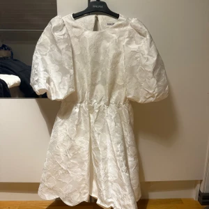 Studentklänning från By Malina - Superfin vit kortklänning från By Malina med puffiga korta ärmar och vackert blommönster i tyget. Klänningen har öppen rygg och markerad midja, vilket ger en riktigt snygg siluett. Perfekt för sommarens alla tillfällen!