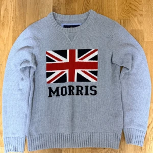 Grå stickad tröja Morris med flagga - Snygg grå stickad tröja från Morris med Union Jack-flagga i rött, vitt och mörkblått på bröstet samt Morris-logga under. Tröjan har rund hals och långa ärmar med ribbade muddar. Perfekt för dig som gillar klassisk stil och vill sticka ut.