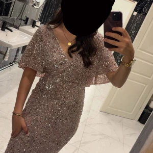 Glittrig champagnefärgad långklänning - Supersnygg långklänning i champagnefärg med massor av glittrande paljetter. Klänningen har v-ringning, omlottliv och vida korta ärmar. Perfekt för dig som vill sticka ut och glänsa på festen! kan användas som en förlovningsklänning