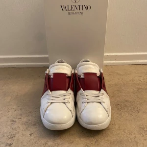 Valentino Garavani Open sneakers vit/röd - Säljer ett par Valentino Garavani Open sneakers i vitt skinn med bred röd läderdetalj över mitten. Klassisk rund tå, vita snören och diskreta nitar på hälen. Snygg och stilren design som sticker ut. Skorna kommer med originalkartong och dustbag. Storlek 40 men passar för 41.