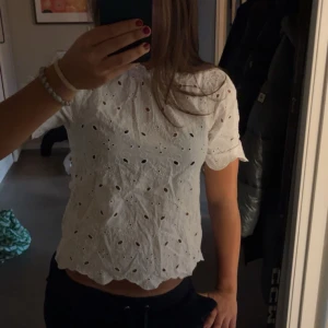 Vit broderad blus Holly & Whyte XS - Superfin vit blus från Holly & Whyte i storlek XS. Blusen har korta ärmar, broderade detaljer och hålmönster över hela tyget. Den har en söt, vågig kant nertill och är gjord i bomull. Perfekt för en fräsch look! 🥰