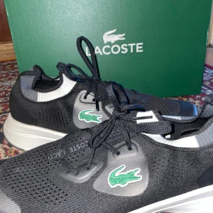 Svarta sneakers från Lacoste - Snygga svarta sneakers från Lacoste med vit och grå detalj vid ankeln och klassisk grön krokodillogga på sidan. Ovandelen är stickad i syntetmaterial för en sportig look och skön passform. Skorna har snörning och vit platt sula som ger en clean vibe. Mer för jogging eller promenad, 42 men väldigt tajta så 41 kan nog också ta på sig dom