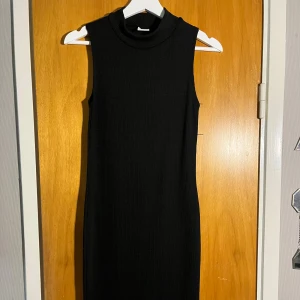 Svart ärmlös ribbad klänning - Snygg svart ärmlös klänning med ribbad struktur och hög krage. Den har en tight passform och är perfekt för dig som gillar minimalistisk stil. Klänningen är kort och passar till många olika tillfällen.