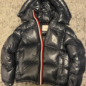 Moncler Montbeliard navy - Säljer en svart, glansig pufferjacka från Moncler med avtagbar huva och klassisk logga på bröstet. Jackan har röda och vita detaljer längs dragkedjan, flera fickor med dragkedja och quiltad design. Perfekt för kalla dagar och riktigt snygg streetstil. Size 5