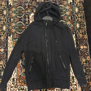 Svart hoodie med goggles från C.P. Company - Svart hoodie från C.P. Company med ikoniska goggles i huvan. Tröjan har dragkedja framtill, två fickor med dragkedja och justerbar huva. Perfekt för dig som vill sticka ut med en streetwear-look.