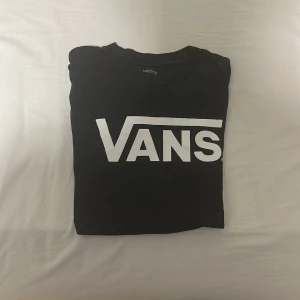 Svart Vans t-shirt med logga - Svart t-shirt från Vans med stor vit logga framtill. Klassisk passform och rund halsringning. Perfekt för dig som gillar streetwear och enkel stil. Tillverkad i mjuk bomull som är skön att ha på sig hela dagen.