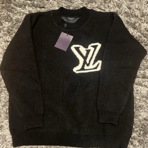 Svart tröja från Louis Vuitton  - Svart stickad tröja från Louis Vuitton i storlek S med stort vitt LV-logo på bröstet. Rund halsringning och ribbade muddar vid ärmslut och nederkant. Perfekt för dig som gillar exklusiva märken och vill ha en stilren look.