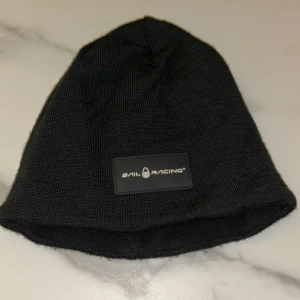 Svart mössa från Sail Racing - Svart mössa från Sail Racing med stilren logotyp framtill. Tillverkad i en mjuk ullblandning som håller dig varm under kyliga dagar. Klassisk passform och enkel design som funkar till de flesta outfits.
