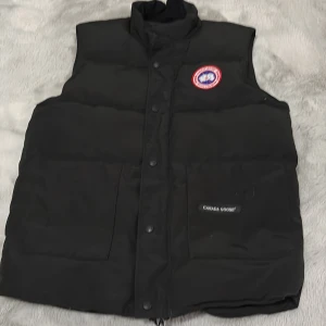 Svart dunväst från Canada Goose - Svart dunväst från Canada Goose med hög krage och tryckknappar framtill. Västen har två stora fickor på framsidan och klassisk Canada Goose-logga på bröstet. Perfekt för kalla dagar och har en stilren, tidlös look.