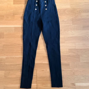 Svarta höga byxor med knappar från Zara - Snygga svarta höga byxor från Zara med sex knappar framtill och veckade detaljer vid midjan. Byxorna har en slim passform och är tillverkade i ett stretchigt material som sitter tajt längs benen. Perfekta för dig som gillar en stilren look med lite edge.