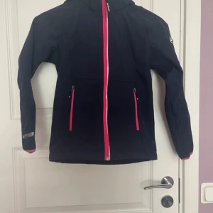 Svart McKinley vindjacka med rosa detaljer - Svart vindjacka från McKinley med snygga rosa dragkedjor och detaljer. Jackan har huva, två fickor med dragkedja och reflekterande band vid ärmslut. Insidan är fodrad med mjuk rosa fleece för extra komfort. Perfekt för dig som gillar en sportig och stilren look.