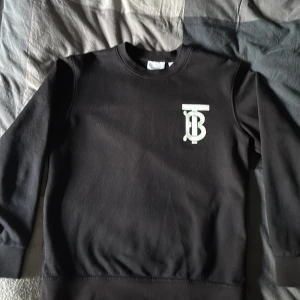 Svart sweatshirt från Burberry - Svart sweatshirt från Burberry med klassisk rund hals och stort vitt TB-logotyptryck på bröstet. Tröjan är långärmad och har ribbade muddar vid ärmslut och nederkant. Perfekt för en clean och stilren look.