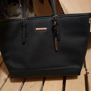 Svart handväska från Tommy Hilfiger - Snygg svart handväska från Tommy Hilfiger med stilren design och guldfärgade detaljer. Väskan har två rejäla handtag, dragkedja upptill och flera innerfack för smart förvaring. Tillverkad i tåligt syntetmaterial med diskret struktur.