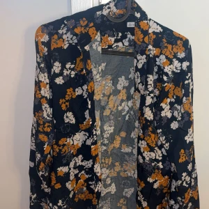 Blommig skjorta från The Shirt Factory - Snygg skjorta med blommigt mönster i vitt, orange och grått på mörkblå botten från The Shirt Factory. Skjortan har lång ärm, klassisk krage och knäppning framtill. Perfekt för dig som vill sticka ut med färg och mönster. Storlek 34. Priset kan diskuteras. Originalpris 1300kr