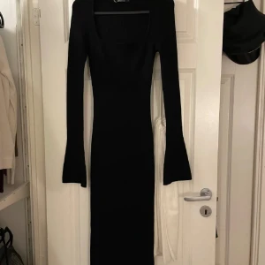 Svart långärmad klänning med fyrkantig ringning - En svart, långärmad klänning med snygg fyrkantig ringning och figurnära passform. Klänningen är lång och har lätt utsvängda ärmslut för en cool siluett. Perfekt för dig som gillar minimalistisk och stilren design.