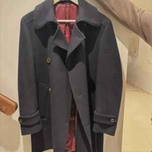 Marinblå kappa från Dressman - Klassisk marinblå kappa från dressman med snygg röd insida. Jackan har dubbelknäppning, stora slag och dekorativa knappar vid ärmsluten. Perfekt för dig som vill ha en tidlös och cool jacka till höst och vinter.