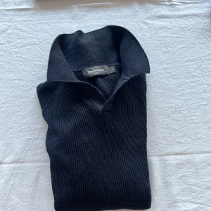 Mörkblå stickad polotröja från Dressman - Klassisk svart stickad polotröja från Dressman med ribbad struktur och hög krage. Tröjan har lång ärm och en stilren, tidlös design som passar perfekt till lager-på-lager. Materialet är mjukt och håller värmen under kyliga dagar.