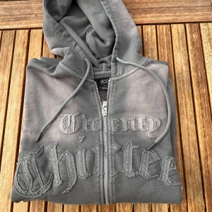 Grå hoodie med broderad text   - Säljer en grå hoodie  med dragkedja och broderad text 'Twenty Thirteen' på bröstet. Tröjan har huva med snörning, två stora fickor fram och en loose passform. Perfekt för dig som gillar streetwear och vill ha en unik look.