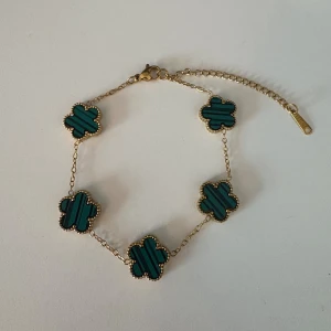 Guldarmband med gröna fyrklöver - Snyggt armband med fem gröna fyrklöverformade hängen och tunn guldfärgad kedja. Klöverhänget har en randig detalj och små pärlkanter runt varje blomma. Perfekt accessoar för att lyfta din stil.