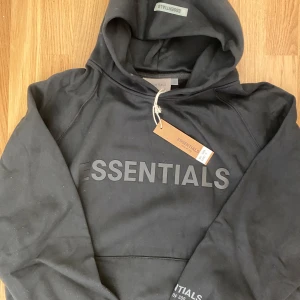 Svart Essentials hoodie med huva - Svart hoodie från Essentials Fear of God med stor logga framtill. Klassisk passform med huva och känguruficka. Tillverkad i mjuk bomullsmix, perfekt för en avslappnad streetwear-look.