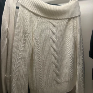 Vit stickad offshoulder-tröja - Säljer en vit stickad tröja med bred offshoulder-krage och snyggt flätmönster. Tröjan har lång ärm och ribbade muddar, perfekt för kyliga dagar. Superfin att styla med jeans eller kjol för en trendig look.