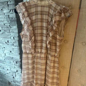 Beige och vit rutig volangklänning - Supersöt kortklänning i beige och vitt med rutigt mönster och volangdetaljer längs axlar och nederkant. Klänningen är ärmlös och har en v-ringning med spetskant upptill. Perfekt för sommarens alla tillfällen!