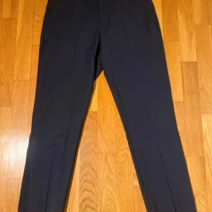 Mörkblå kostymset J.Lindeberg  - Snygga mörkblå kostymset från J.Lindeberg i storlek 44. Byxorna har klassisk rak passform med pressveck och dragkedja i gylfen. Perfekta till en dressad look och tillverkade i ett mjukt material med lite stretch för extra komfort. Använd 1 gång