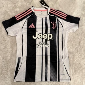 Juventus fotbollströja Adidas svart/vit - Juventus matchtröja från Adidas med klassiska svarta och vita ränder, rosa detaljer på axlar och logga. Jeep som sponsor på bröstet och 'Visit Detroit' tryck. Kortärmad, tillverkad i lätt och ventilerande polyester med Aeroready-teknologi.