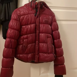 Vinröd pufferjacka från XP-D - Säljer en vinröd pufferjacka från XP-D med huva. Jackan är väl använd och har tappat båda knapparna vid fickorna, på baksidan är det som lite sträck på jackan men inget som är mer sönder, säljer den billigt 