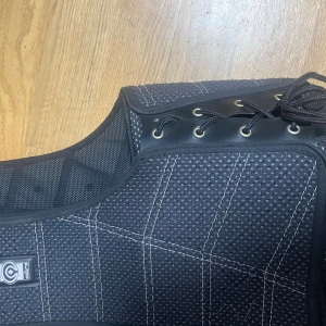 Svart Champion Cobra ridväst M - Svart skyddsväst från Champion Cobra med vit kontrastsöm, snörning i sidorna och dragkedja framtill. Västen är tillverkad i slitstarkt syntetmaterial och har meshpaneler för extra ventilation. Perfekt för dig som rider och vill ha extra säkerhet.