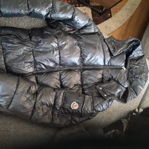 Moncler Maya - Säljer en svart pufferjacka från Moncler med glansig finish, huva och dubbla dragkedjor. Jackan har Moncler-logga på ärmen och klassisk quiltad design. Perfekt för kalla dagar och riktigt snygg streetstil. Passade inte mig så säljer den för att köpa en annan storlek! 
