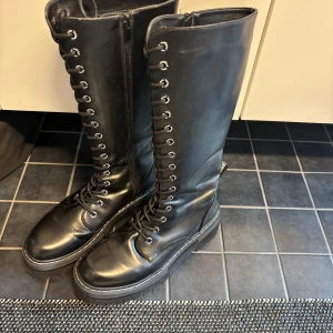 Svarta höga snörade stövlar/kängor från Dockers  - Säljer ett par svarta höga stövlar med snörning hela vägen upp och dragkedja på insidan. De har rund tå, platt sula och är tillverkade i blankt skinn. Perfekt för dig som gillar en edgy stil och vill ha boots som sticker ut. Sparsamt använda.
