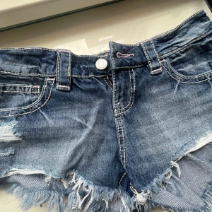 Blå jeansshorts med fransig kant - Säljer ett par blå jeansshorts med låg midja och rå, fransig kant. Shortsen har slitna detaljer, klassiska fickor fram och bak samt vita kontrastsömmar. Från Gina Tricot 14+ collection, helt nya inte använda en ända gång för jag beställde i fel storlek men tog bort lappen 🫢midja= 39 cm. Längd= 27cm 