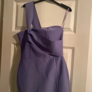 Lila asymmetrisk klänning med slits - Säljer en lila figurnära klänning med asymmetrisk axel och en snygg slits framtill. Klänningen har en axelbandslös sida och en bred axelrem, vilket ger en cool och modern look. Materialet är slätt och stretchigt, perfekt för dig som vill sticka ut.