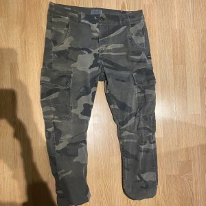 Camouflage cargopants från Jack & Jones - Säljer ett par snygga cargopants från Jack & Jones med klassiskt camouflage-mönster i gröna och grå toner. Byxorna har flera praktiska fickor med knappstängning och är tillverkade i slitstark bomull. Perfekt för dig som gillar streetwear och funktionella detaljer.