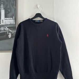 Svart sweatshirt från Polo Ralph Lauren - Svart sweatshirt från Polo Ralph Lauren med klassisk röd broderad logga på bröstet. Tröjan har rund halsringning, långa ärmar och ribbade muddar. Perfekt för en clean och stilren look.