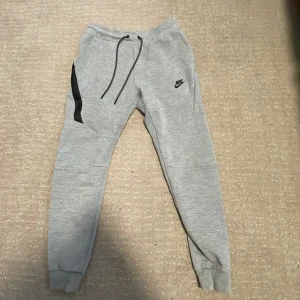 Nike tech old version  - Grå mjukisbyxor från Nike med svart logga på vänster lår och svart snörning i midjan. Byxorna har en svart detalj längs ena benet och muddar vid bensluten. Perfekta för chill eller träning, riktigt sköna och stilrena.
