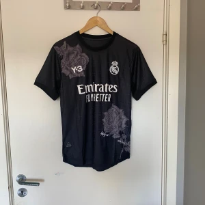 Real Madrid x Y-3 svart fotbollströja M - Snygg svart Real Madrid fotbollströja i samarbete med Y-3 och Yohji Yamamoto. Tröjan har korta ärmar, vita tryck och unika blommotiv i grått. Slim fit-modell i lätt och ventilerande material, perfekt för dig som vill sticka ut på planen eller gatan.