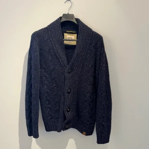 Mörkblå stickad kofta med knappar - Mysig mörkblå stickad kofta med grov ribbad krage och snygga knappar framtill. Koftan har kabelstickat mönster på ärmarna och är långärmad, perfekt för kyliga dagar. Passar dig som gillar klassisk och stilren look. Den är i ull och har inga defekter, hör av er vid frågor eller fler bilder