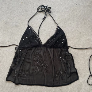 Svart mesh linnetopp med paljetter - Säljer en svart mesh linnetopp med tunna knytband i nacken och på sidorna. Toppen har v-ringad front och är dekorerad med glittriga paljetter och pärlor i blommönster. Materialet är transparent och ger en lätt och luftig känsla. Liite trasig på kanten men syns ej