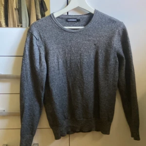 Grå stickad tröja från J.Lindeberg - Säljer en stilren grå stickad tröja från J.Lindeberg. Tröjan har rund halsringning och långa ärmar, perfekt för lager-på-lager. Enkel och klassisk design som funkar till det mesta. Snyggt melerad yta och mjukt material.