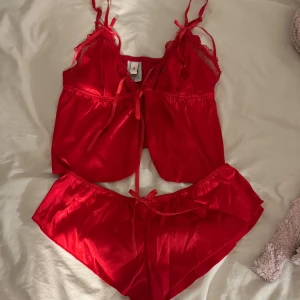 Röd satin linnetopp från Nelly samt underdel - Säljer en röd linnetopp från Nelly i glansig satin med spetsdetaljer och smala axelband. Toppen har en söt rosett framtill och är croppad i modellen. Perfekt för dig som vill ha något extra till myskvällen. Det följer även med en underdel. 