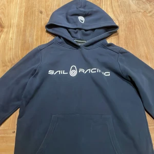 Marinblå Sail Racing hoodie - Snygg marinblå hoodie från Sail Racing med vit logga tryckt på bröstet. Tröjan har huva, lång ärm och en stor magficka framtill. Perfekt för dig som gillar sportig och avslappnad stil. Pris kan diskuteras