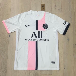PSG vit och rosa fotbollströja Nike - Snygg PSG fotbollströja från Nike i vit med en rosa och svart vertikal rand på framsidan. Kortärmad modell med svarta detaljer på ärmslut och klubbmärke på bröstet. Materialet är lätt och andas, perfekt för träning eller match.