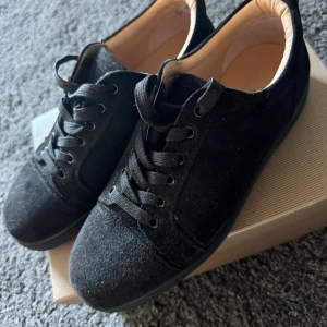 Svarta sneakers från Christian Louboutin - Snygga svarta sneakers från Christian Louboutin i mocka med klassisk röd sula och svarta skosnören. Skorna har rund tå och platt sula, samt lyxig innersula i beige läder. Perfekta för dig som vill sticka ut med exklusiva detaljer. Skorna är i storlek 43 och passar perfekt till alla årstider om du vill sticka ut! Hör av er vid fler frågor.